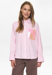 Roze en witte gestreepte blouse met lange mouwen en een zak met paillettenaccent. De stof heeft een gladde textuur; de kraag staat rechtop.