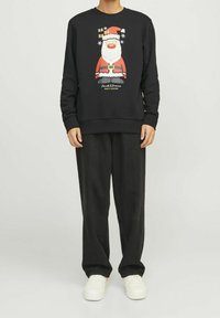 Una persona indossa un felpa nera con una grafica colorata di Babbo Natale, abbinata a pantaloni neri larghi e sneakers bianche.