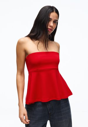 Woman with long dark hair wearing a strapless red peplum top and dark jeans, standing against a plain white background.