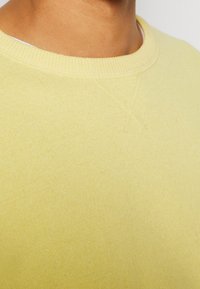 Heller gelber Pullover mit geripptem Rundhalsausschnitt und glatter Textur; verfügt über ein kleines Kreuzstichdetail nahe dem Kragen.