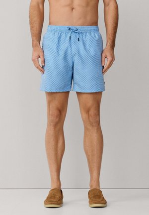 Mann trägt hellblaue, gemusterte Badeshorts mit schwarzem Kordelzug und braune Slipper, steht vor einfarbigem Hintergrund.