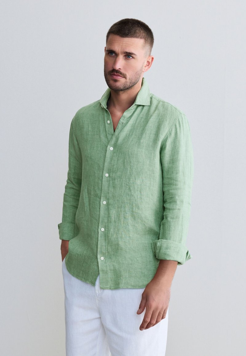 Chemise en lin verte à col classique, manches longues et boutonnée à l'avant. Portée avec un pantalon en lin blanc, mettant en valeur une coupe décontractée et une texture.