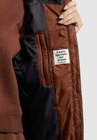 Braune gesteppte Innenjacke mit marinefarbener Fütterung. Verfügt über ein gewebtes Etikett mit dem Text "Eine warme Umarmung aus Stoff." Weicher Stoff und saubere Nähte.