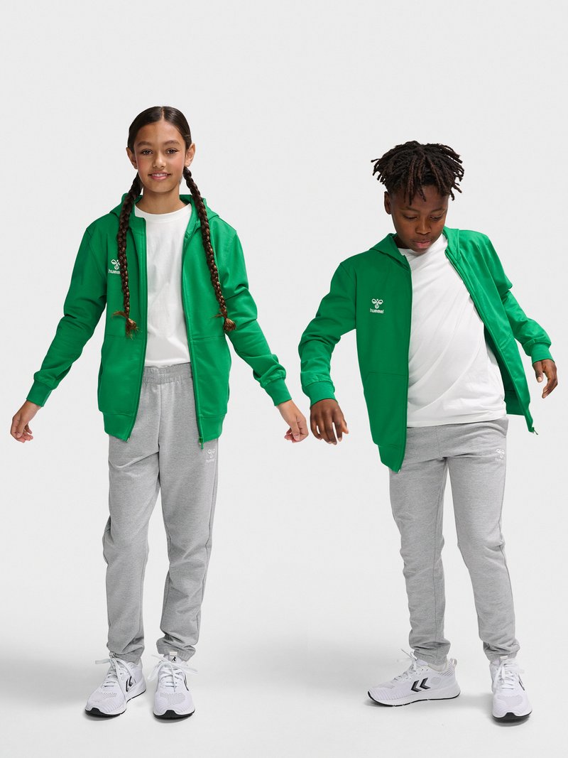 Deux enfants portant des sweats à capuche verts zippés, des chemises blanches, des pantalons de jogging gris et des baskets blanches se tiennent devant un fond uni.