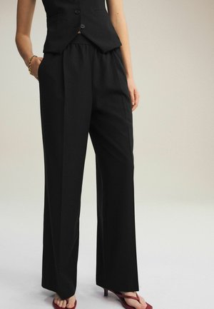 Schwarze maßgeschneiderte Hose mit entspanntem Sitz, ausgestattet mit einem elastischen Bund und einer glatten Textur, ergänzt durch offene rote Absatzsandalen.