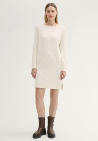 Cremfarbendes gestricktes Kleid mit Zopfmuster, gerippten Bündchen und Saum. Mit seitlichen Schlitzen und einer lässigen Silhouette. Kombiniert mit braunen Ankle Boots.