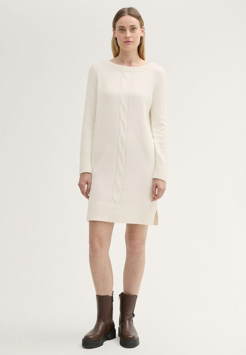 Cremfarbendes gestricktes Kleid mit Zopfmuster, gerippten Bündchen und Saum. Mit seitlichen Schlitzen und einer lässigen Silhouette. Kombiniert mit braunen Ankle Boots.