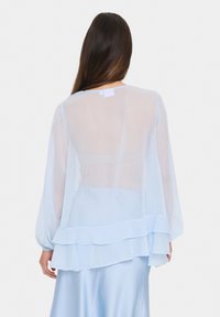 Blusa trasparente azzurro chiaro con maniche lunghe, orlo a strati e tessuto leggermente strutturato. La vista posteriore mostra un collo pulito e senza chiusure visibili.