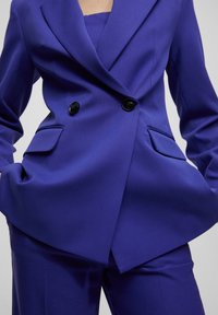 Veste blazer violette sur mesure avec boutons croisés, revers crantés et poches latérales, assortie à un pantalon coordonné. Texture lisse.