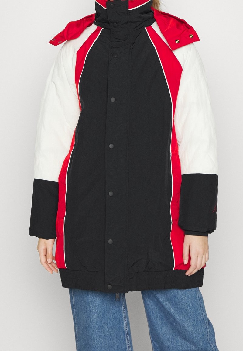 Person iført en lang vinterjakke med farveblokke i rød, sort og hvid, kombineret med blå jeans, stående mod en ensfarvet baggrund.