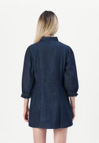 Kvinna med långt blont hår som bär en mörkblå denim minikjol med trekvarts puffärmar, sedd bakifrån mot en vit bakgrund.