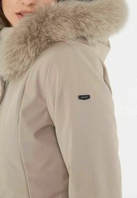 Beżowa zimowa kurtka z kapturem z sztucznego futra, gładka tekstura materiału oraz mały czarny detal z logo na rękawie.
