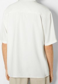 Camicia a maniche corte con colletto in tessuto crema chiaro, caratterizzata da una texture liscia, un fit leggermente più ampio e un sottile dettaglio a yoke sul retro.