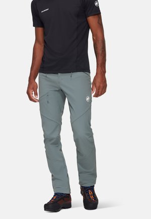 Pantaloni da trekking grigio chiaro con un design slim, tasche laterali e logo; abbinati a una maglietta nera a maniche corte e scarpe nere resistenti.