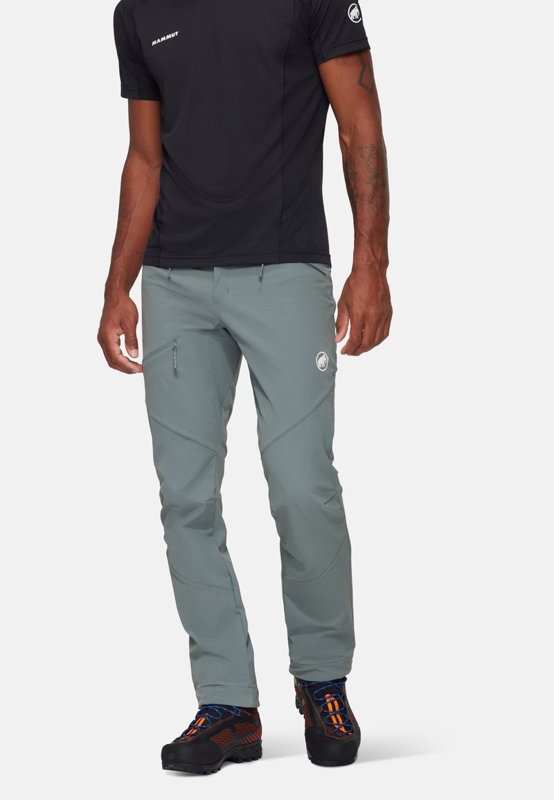 Pantalons de randonnée gris clair avec un design ajusté, des poches latérales et un logo ; associés à un t-shirt noir à manches courtes et des chaussures noires robustes.