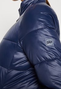 Kobieta w błyszczącej granatowej kurtce puchowej z wysokim kołnierzem i logo GAP na górnej części rękawa, pokazana z boku.