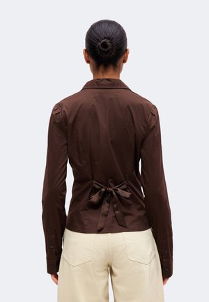 Vue de dos d’une femme portant un haut marron à manches longues noué à la taille et un pantalon crème, sur un fond clair uni.