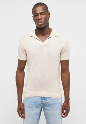 Mann trägt ein cremefarbenes kurzärmeliges Strick-Poloshirt und hellblaue Jeans, steht vor einem schlichten hellen Hintergrund.