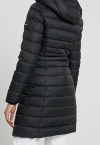 Parka acolchada negra con capucha, presenta patrones acolchados horizontales y un ligero ajuste en la cintura. Incluye detalle de logo en la manga.