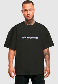 Tricou negru supradimensionat cu mâneci scurte, având un logo holografic "BY STUDIOS" pe piept. Textură moale din bumbac.
