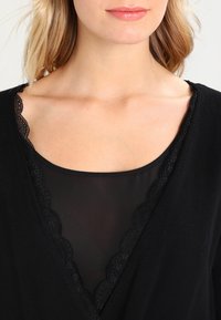 Czarna bluzka z falbaniastym koronkowym wykończeniem przy dekolcie, nałożona na przezroczystą, czarną tkaninę. Gładka faktura i dekolt w serek.