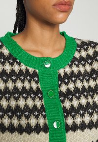 Pletený kardigan s zeleným žebrovaným límcem a knoflíky, se vzorem houndstooth v černé a krémové barvě. Měkká textura a kontrastní akcenty.