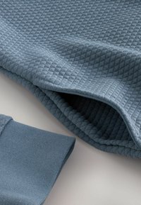 Un tejido azul texturizado presenta un patrón de diamantes con puños acanalados, mostrando una textura suave y tejida y un tono ligeramente más claro cerca de los bordes.