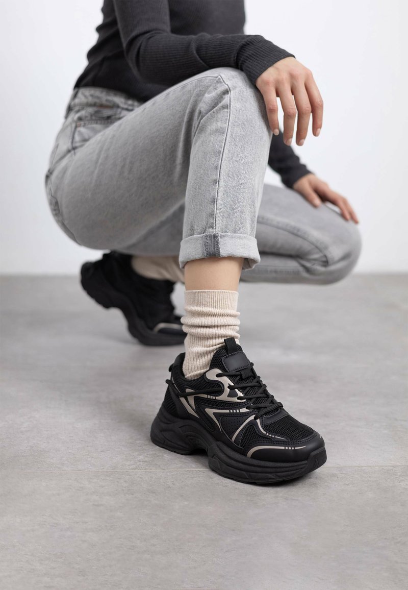 Persona con jeans gris claro y parte superior negra en cuclillas, que lleva zapatillas negras y beige con calcetines color crema sobre un suelo gris.