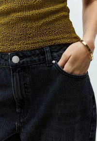 Schwarze Denim-Jeans mit hoher Taille, ausgestattet mit einem silbernen Knopf und Nieten. Das Oberteil besteht aus strukturiertem Senfstoff, der die Taille betont.