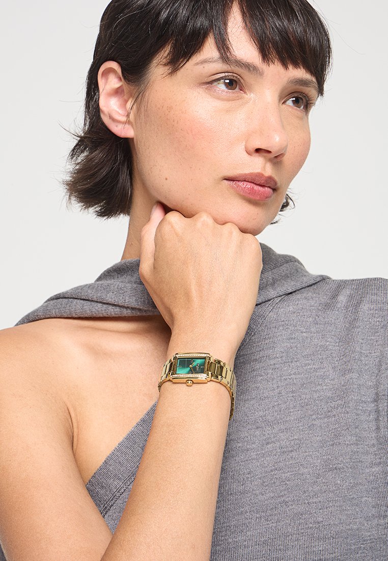 Goldfarbene rechteckige Uhr mit grünem Ziffernblatt, Gliederarmband und dekorativen Akzenten. Am Handgelenk getragen, mit einem strukturierten grauen Kleidungsstück.