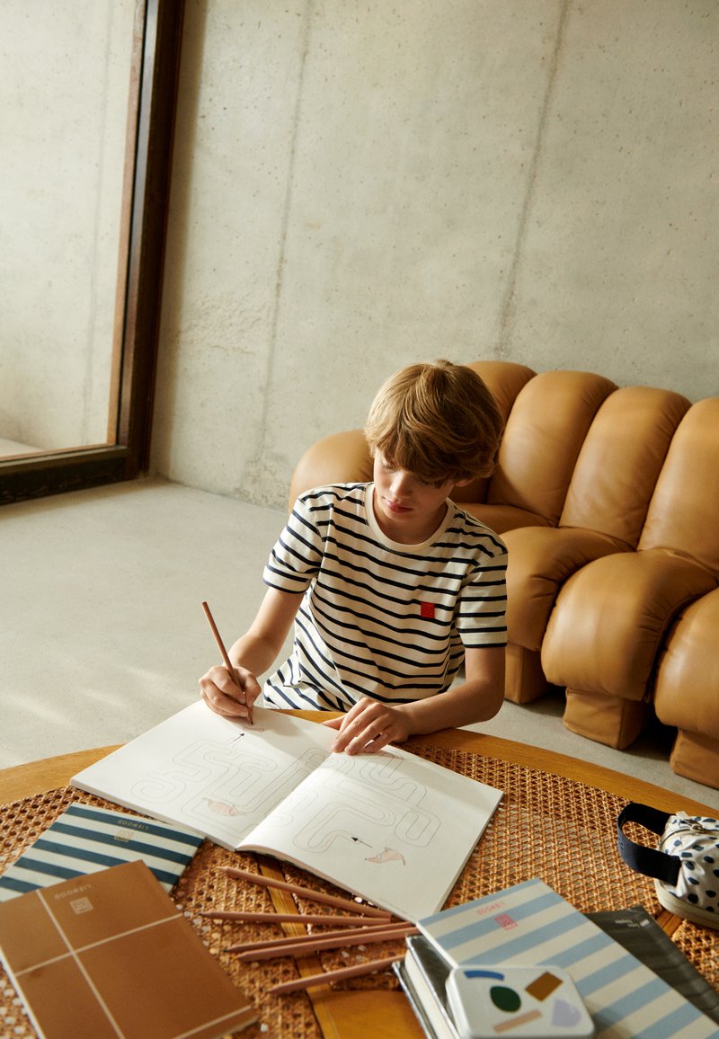 Striped t-shirt with red accent, seated at a wooden table with a woven surface, coloring in a sketchbook. Soft brown leather sofa in background.