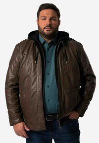 Chaqueta de cuero marrón con capucha negra, con cierre frontal, bolsillos y detalles en los hombros. Llevada sobre una camisa de botones en tono verde azulado.