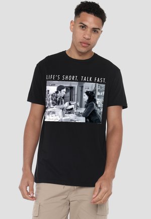 Young man wearing black T-shirt with black-and-white scene of two people talking at a kitchen counter and caption "Life’s short. Talk fast."
