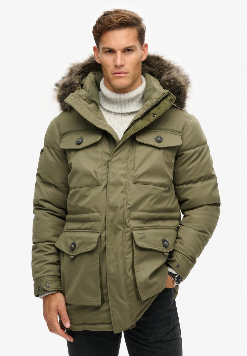 Chaqueta de plumas verde oliva con capucha forrada de piel, múltiples bolsillos frontales y cierres de botones. Tejido texturizado con un acabado suave.