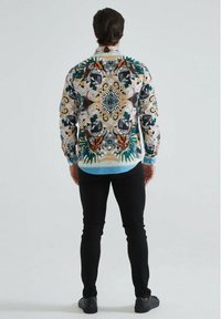 Chemise à manches longues avec un motif florale et animal multicolore vif, avec un col et des poignets bleu clair. Portée avec un pantalon noir.