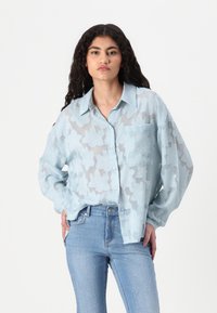 Camisa azul clara, transparente, com botão e textura floral, mangas longas e bolso no peito, combinada com jeans azul claro com adornos.