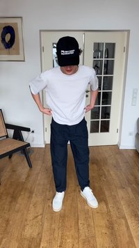 Νεαρός άνδρας με λευκό μπλουζάκι, σκούρο μπλε παντελόνι, λευκά αθλητικά παπούτσια και μαύρο καπέλο στέκεται σε εσωτερικό χώρο σε ξύλινο πάτωμα με γυάλινες πόρτες πίσω του.