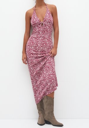 Femme portant une robe midi rouge à bretelles avec un imprimé floral blanc et des bottes de cowboy en suède beige mi-mollet, se tenant contre un fond blanc.