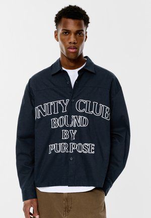 Námořnická modrá košile na knoflíky vyrobená z texturované látky, s velkým bílým vyšíváním, které říká "UNITY CLUB BOUND BY PURPOSE."