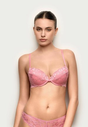 Giovane donna con capelli tirati indietro, che indossa un reggiseno di pizzo rosa e mutandine abbinate, in piedi contro uno sfondo chiaro e semplice.