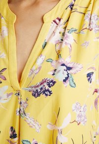 Gelbe Blumenshirt mit V-Ausschnitt, leichtem Stoff und einer Mischung aus lila und grünen Blumenmustern. Elegante Nahtdetails sind vorhanden.
