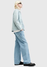 Chaqueta de mezclilla azul clara y jeans de pierna ancha con un acabado texturizado, combinados con zapatos negros y gafas de sol oscuras. Fondo neutro.