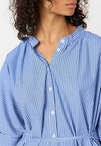 Donna con capelli ricci scuri che indossa un abito camicia a righe verticali blu e bianche con una sottile cintura abbinata.