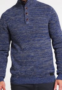 Blauer Strickpullover mit hohem Kragen, vier braunen Knöpfen, einem strukturierten Muster und einer schmalen Passform. Schwarzes Logo-Patch am Saum.