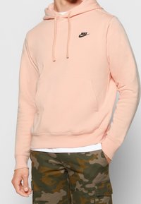 Ljusrosa huvtröja i mjukt tyg, med framficka och svart Nike-logga på bröstet. Matchas med camouflagemönstrade cargobyxor.