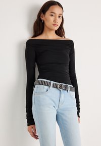 Zwarte off-shoulder top met lange mouwen en geplooide details, gecombineerd met lichtblauwe spijkerbroek en een zwarte riem met studs en een zilveren gesp.