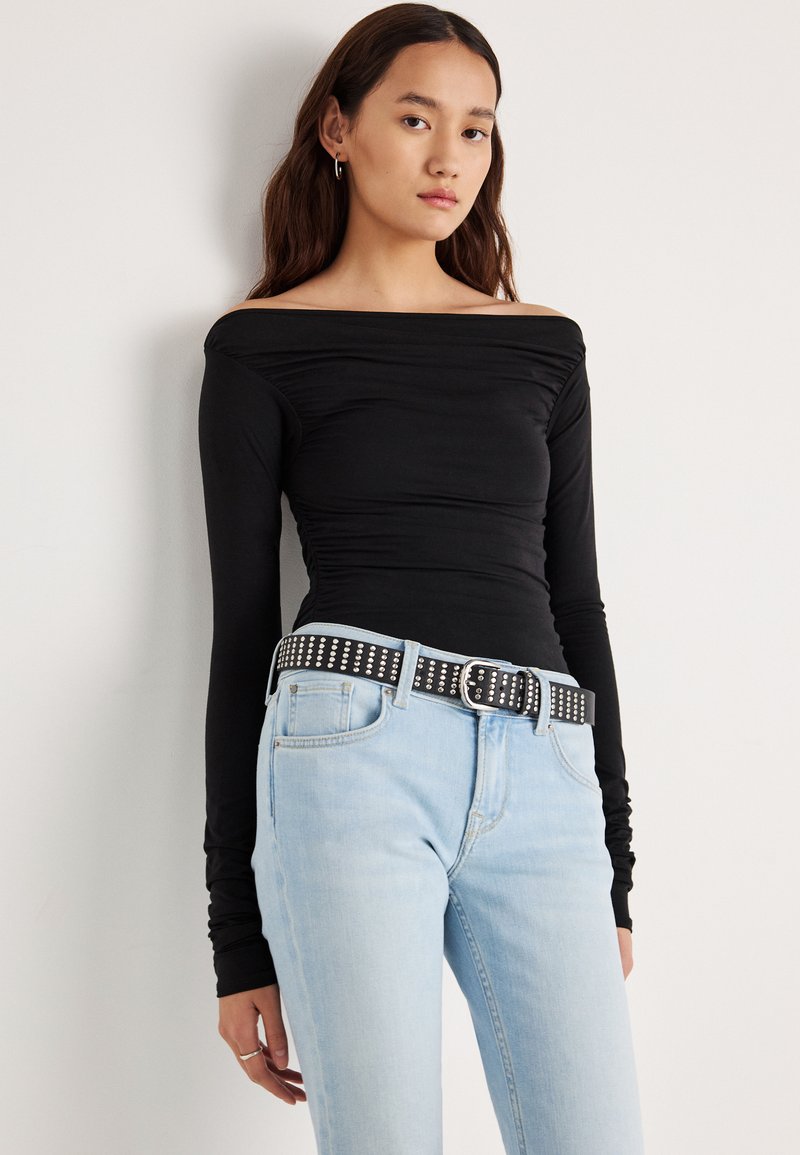Zwarte off-shoulder top met lange mouwen en geplooide details, gecombineerd met lichtblauwe spijkerbroek en een zwarte riem met studs en een zilveren gesp.