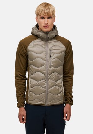 Uomo biondo che indossa una giacca trapuntata con cappuccio marrone e beige con tasca con cerniera sul petto e pantaloni scuri, rivolto verso il davanti.