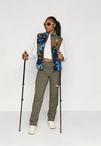 Fleeceväst med flerfärgat blått och brunt mönster, vit långärmad tröja, olivgröna cargobyxor och vandringsstavar; neutrala sneakers.