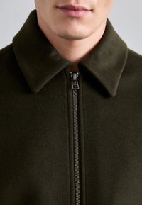 Tamsiai žalia vilnos striukė su plačia apykakle ir sidabrinėmis užtrauktuko detalėmis priekyje. Minkšta tekstūra su elegantišku dizainu.